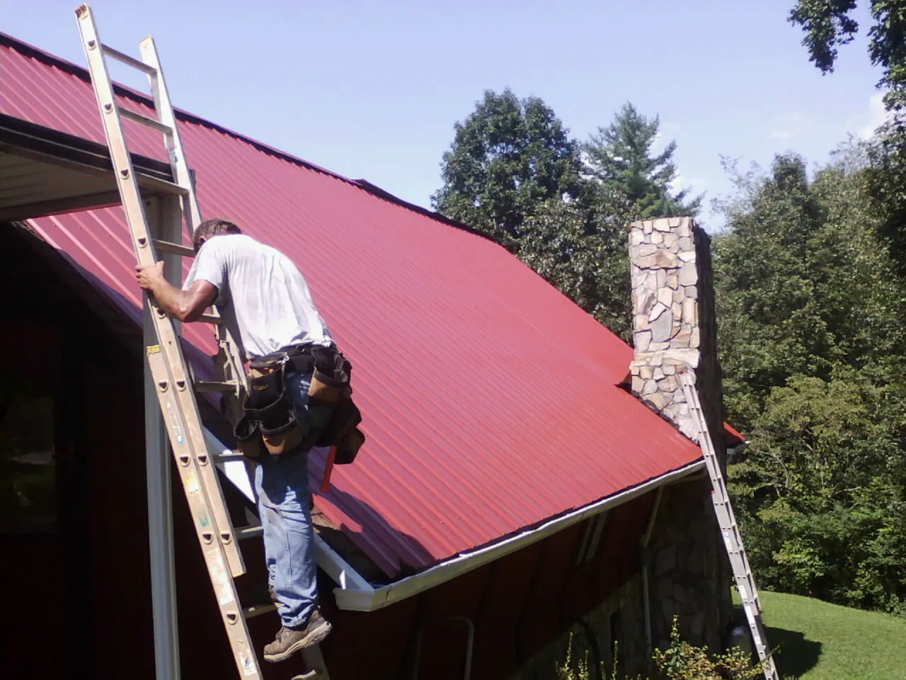 Roofer on a ladder beside a red metal roof and stone chimney — Soffit & Fascia Repair by Roofing Brothers in Hershey